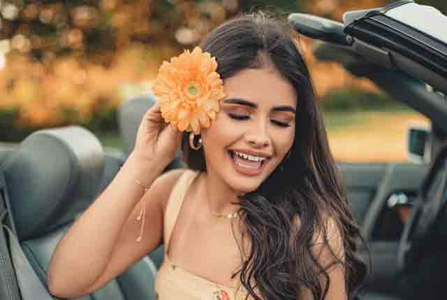 A beautiful, smiling Colombian woman sitting in the passenger seat of a car while holding a flower up against her right ear