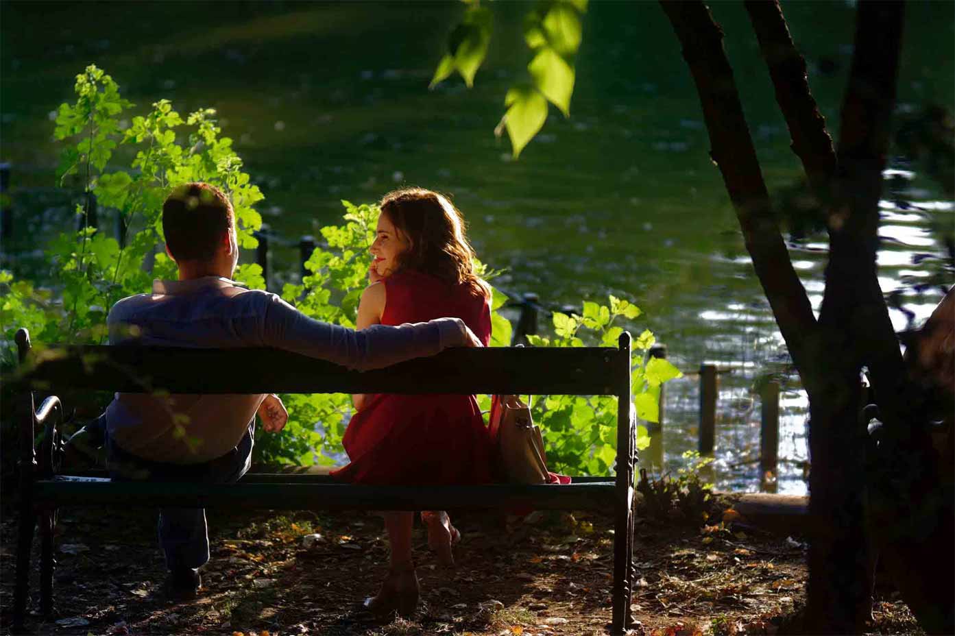 A photo of a couple looking at each other while sitting on a bench facing the lake