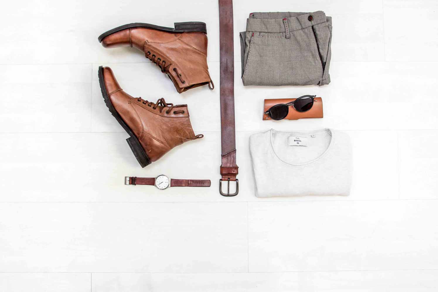 A layout photo of a pair of brown leather boots arranged alongside a brown belt, a watch, a pair of sunglasses, as well as a neatly folded pair of dress pants and a white shirt